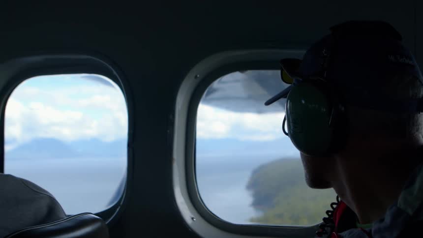 A man wearing headphones looks out the window of a small airplane, taking in the stunning aerial view of a forested coastline