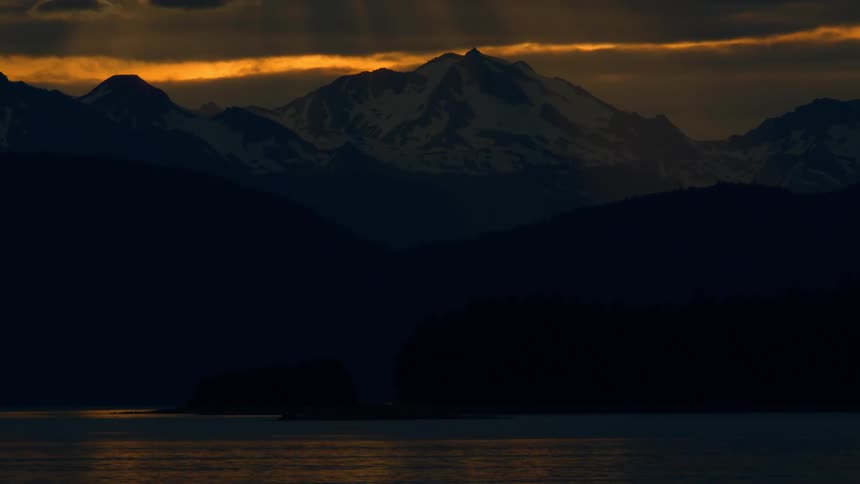 A dramatic sunset casts golden light and sun rays over a majestic snow-capped mountain range above a tranquil body of water