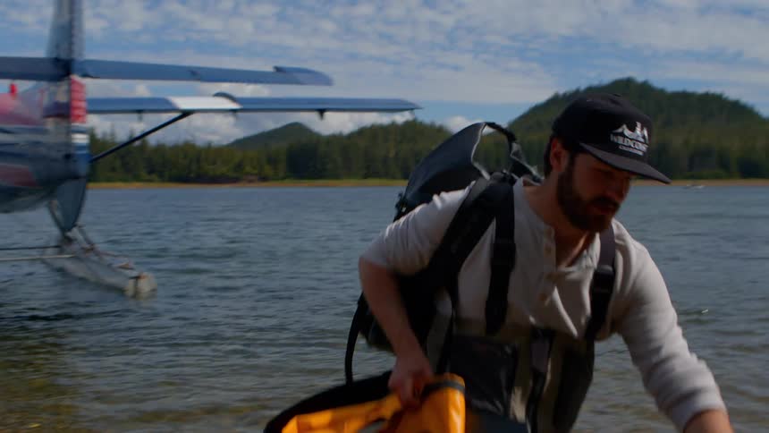 An adventurer carrying a large backpack and gear walks ashore after arriving by seaplane to a remote wilderness lake