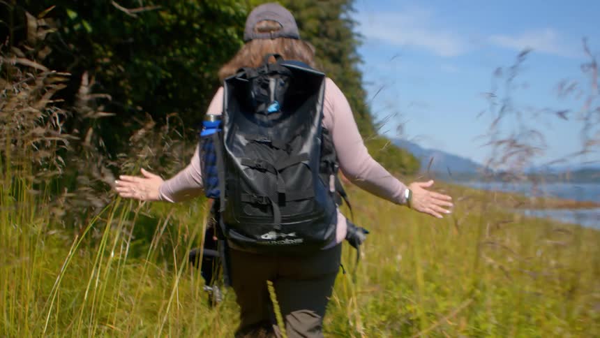 A woman with her arms outstretched hikes through tall grass along a coastal trail with friends in slow motion