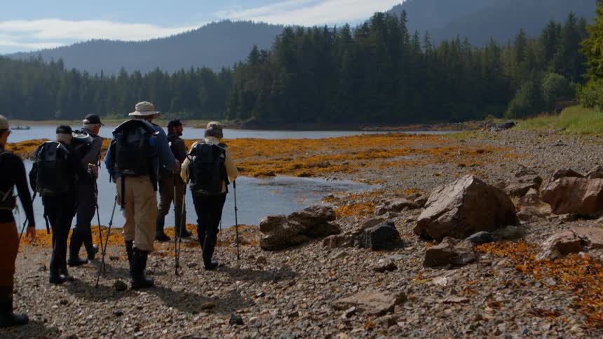 A guided hiking tour group with backpacks and trekking poles walks along a rocky shoreline in a remote wilderness
