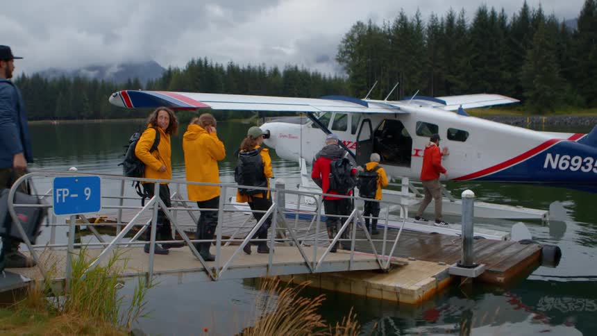 A group of hikers in rain gear walks down a gangway to board a seaplane docked on a calm lake surrounded by an evergreen forest