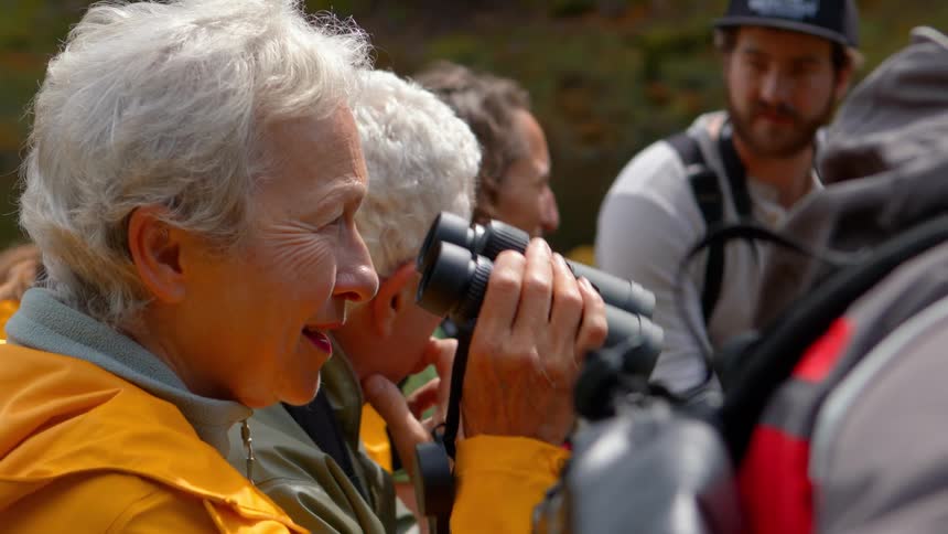 A senior woman in a yellow jacket smiles as she raises her binoculars to get a better view while on a nature tour with a group