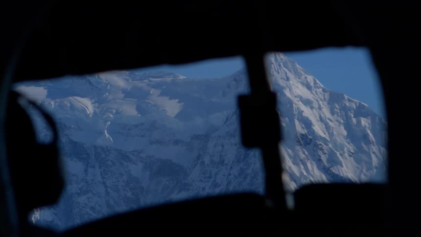 An aerial point-of-view from inside a cockpit reveals a majestic, snow-covered mountain peak against a clear blue sky