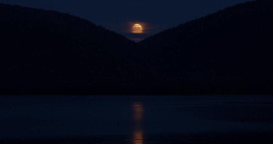 A golden full moon rises between two silhouetted mountains, its light reflecting on the serene lake at night