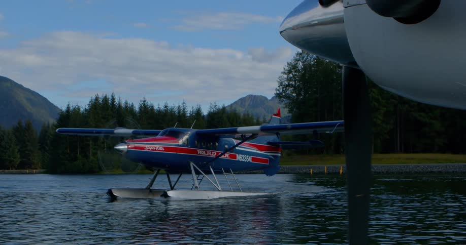 A seaplane's propeller spins in the foreground as another seaplane taxis across a tranquil lake surrounded by a dense forest and mountains