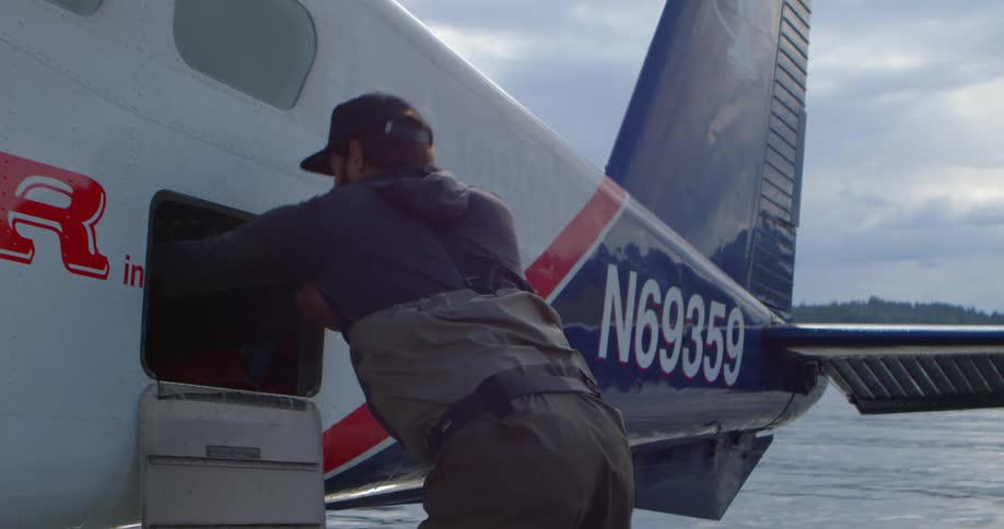 An adventurous man in fishing waders unloads a large backpack from a seaplane onto a wooden dock