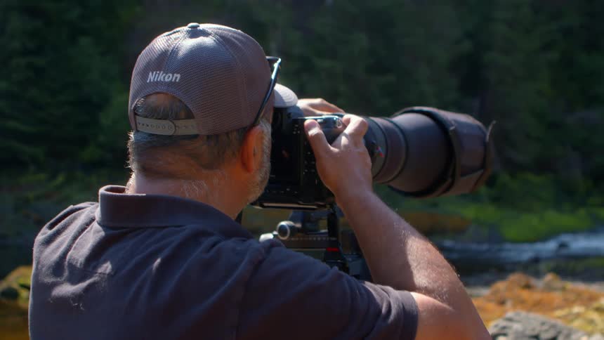 Over-the-shoulder shot of a nature photographer adjusting the settings on his telephoto lens camera mounted on a tripod