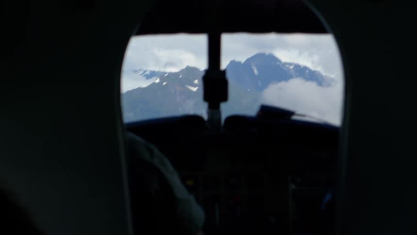 A shaky view from inside a silhouetted cockpit shows a small airplane flying towards snow-capped mountains