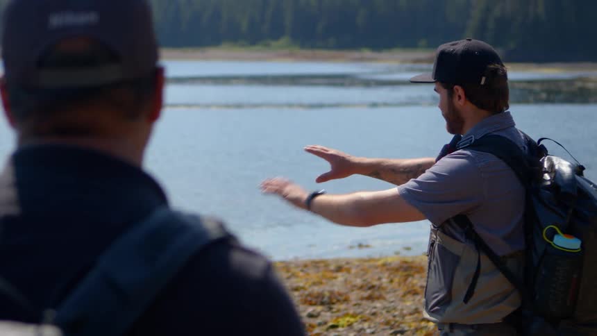 An expert guide gestures towards the water while explaining the scenic area to two hikers on a rocky shore