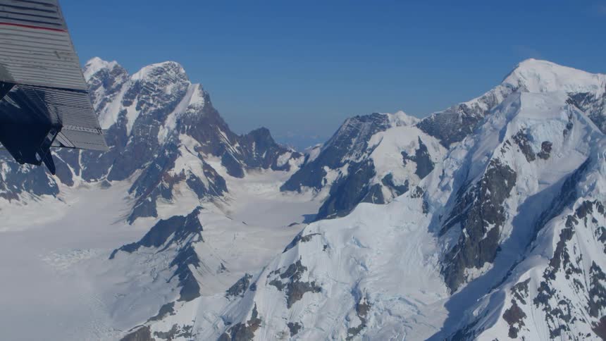 An airplane flies over a vast and majestic snow-covered mountain range on a clear sunny day