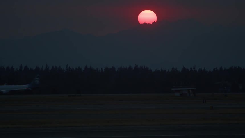 A commercial jet moves across the tarmac under a dramatic, hazy red sunset over a mountain range