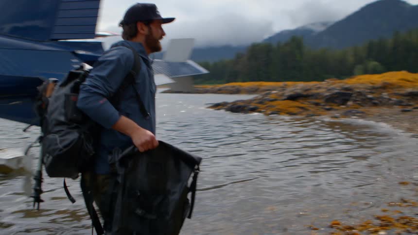 A guide carries his gear ashore after disembarking from a seaplane, joining his tour group on a remote Alaskan coastline