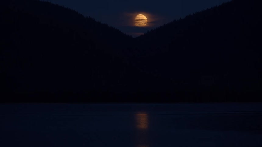 A full moon rises between two silhouetted mountains at night, its golden light reflecting on the calm water below