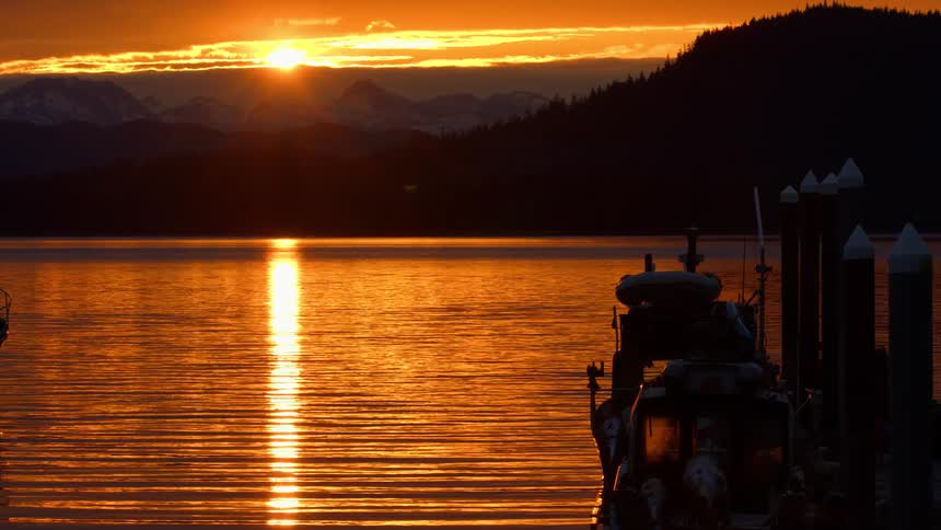A stunning golden sunset reflects on the calm waters of a mountain lake, with fishing boats silhouetted at the dock