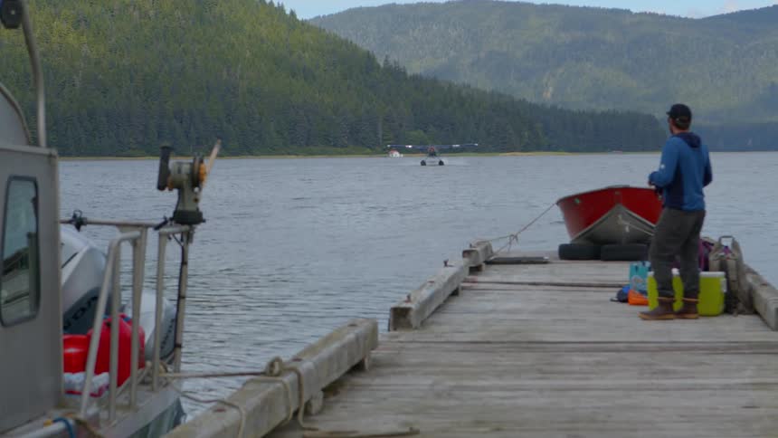A man on a wooden dock watches a seaplane come in for a landing on a remote mountain lake
