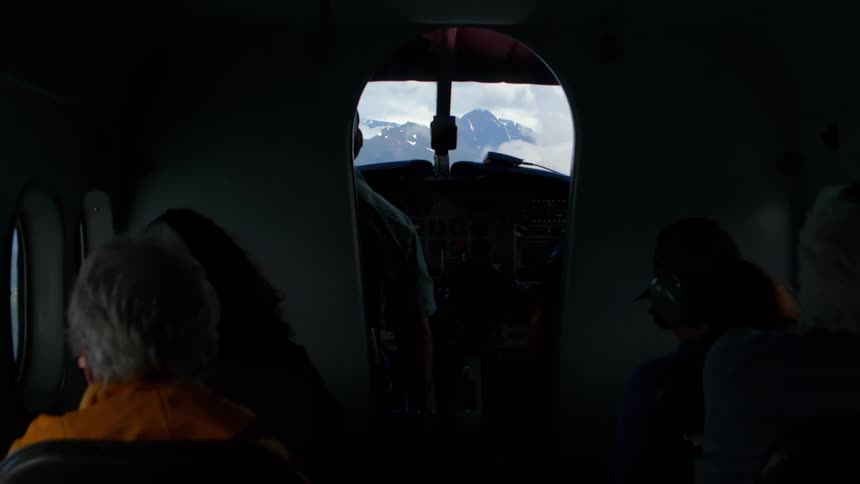 A view from inside a small airplane shows passengers looking towards the cockpit at snow-capped mountains in the distance