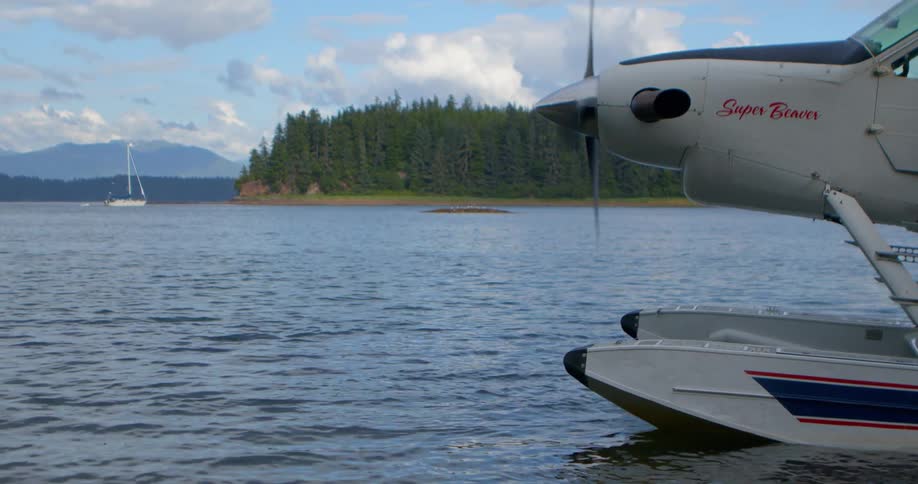 A seaplane with its propeller spinning sits docked on the water with a sailboat and forested coastline in the background