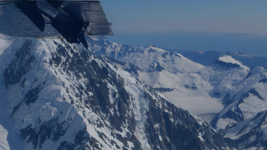 An aerial view from under a small plane's wing reveals a vast and majestic snow-covered mountain range on a clear day