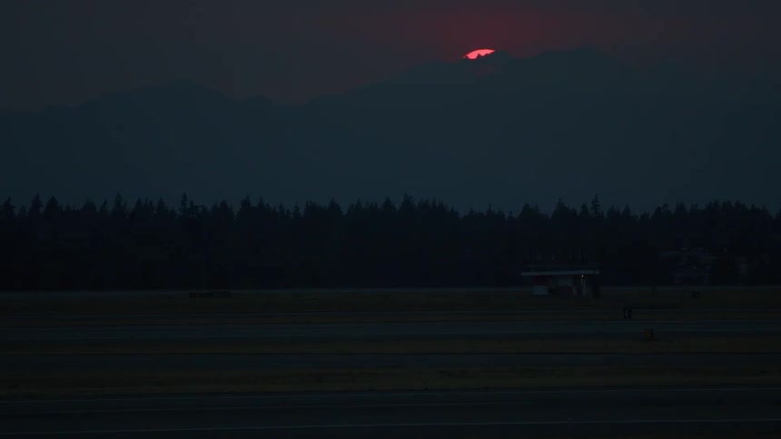 Time-lapse of a hazy red sun setting behind a mountain range over an empty airport runway at dusk