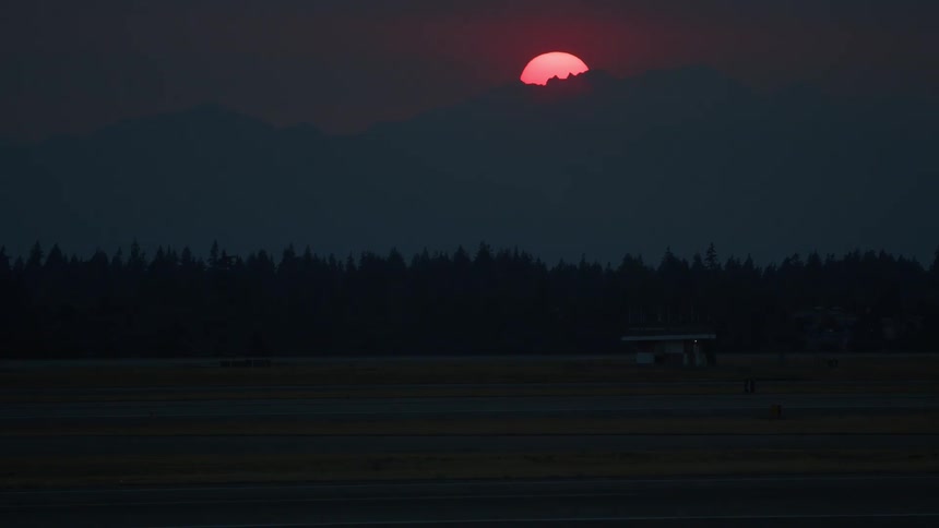 A vibrant red sun dips behind a dark mountain silhouette, casting an eerie glow over an empty airport runway at dusk