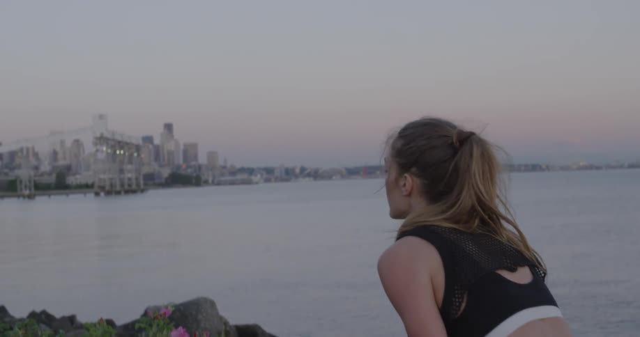 A woman stands next to a body of water with city buildings in the distance