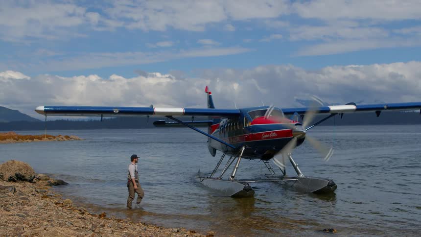 A floatplane with a spinning propeller taxis to a rocky shore where a man in waders climbs aboard