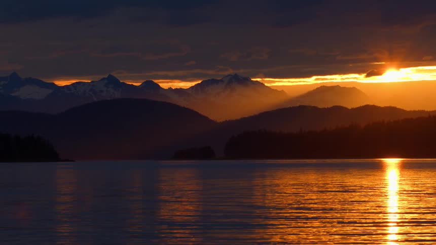 A dramatic golden sunset reflects on a calm lake as sun rays illuminate distant snow-capped mountains