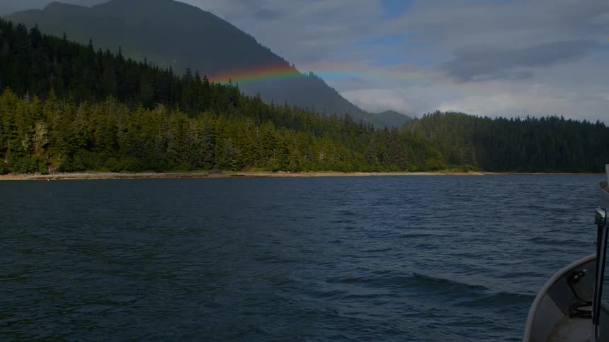A vibrant rainbow appears in the cloudy sky above a lush green, tree-covered mountain and a calm body of water