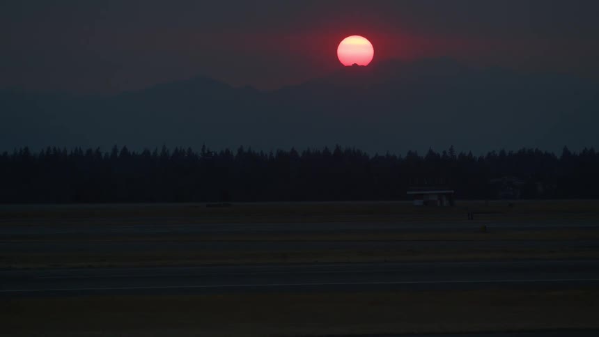 A vibrant red sun sets behind a mountain range, casting a hazy glow over a dark and empty airport runway