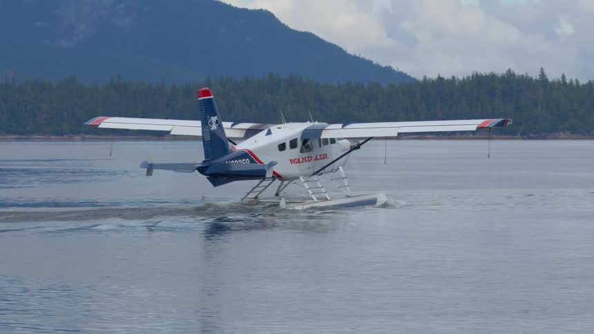 A Ward Air seaplane taxis across a calm lake, preparing for takeoff against a backdrop of forested mountains in Alaska