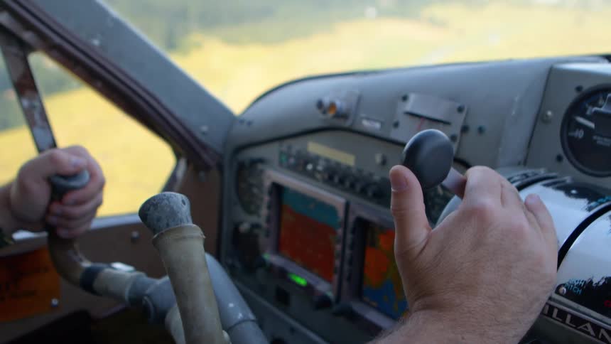 A close-up, shaky shot from inside a small airplane cockpit reveals the pilot's hands on the yoke and throttle controls during flight