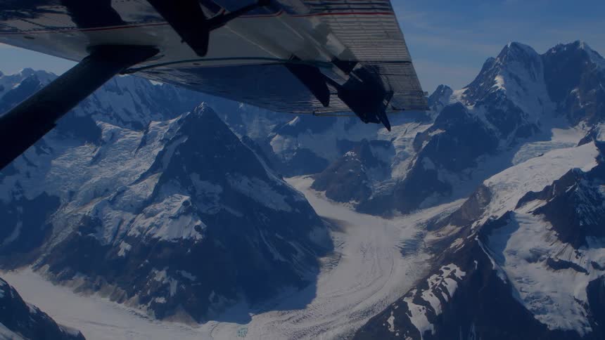 Aerial view from under a plane's wing flying over a vast, snow-covered mountain range and glaciers on a clear day