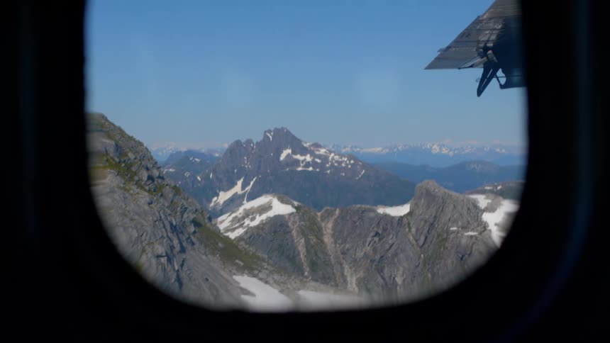 An aerial view of a rugged, snowy mountain range as seen through the porthole of a small aircraft on a sunny day