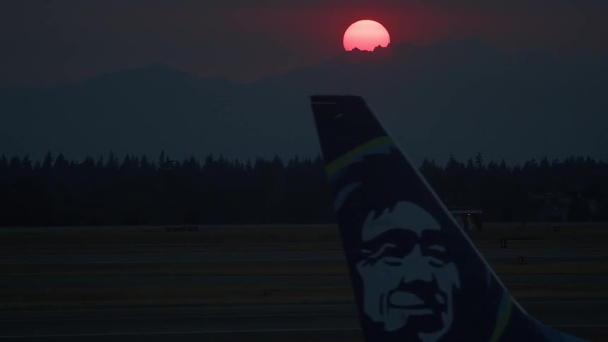An Alaska Airlines airplane taxis on the tarmac during a dramatic, smoky red sunset over a mountain range