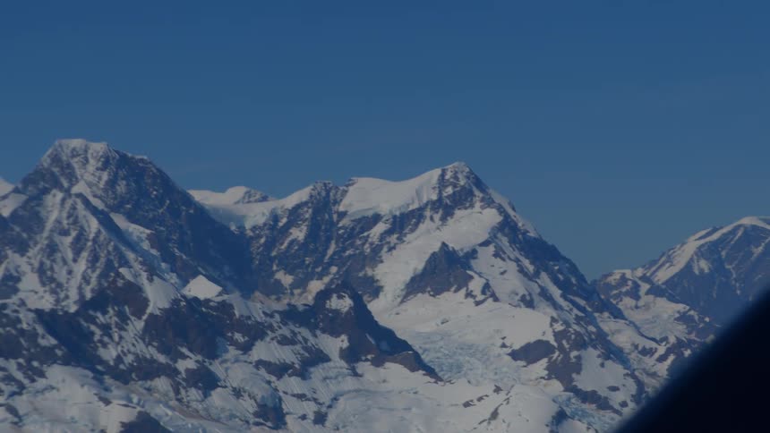 A slow aerial zoom-out reveals the vast expanse of a snow-covered mountain range under a clear blue sky