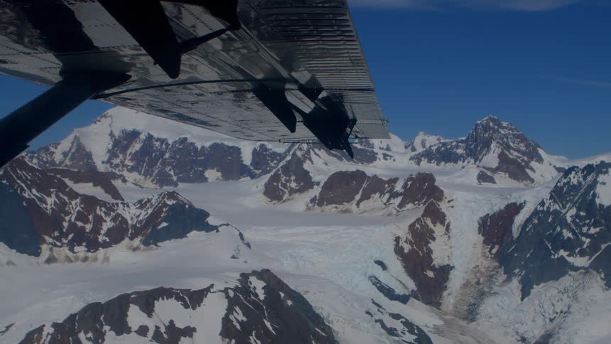 An aerial view from an airplane wing shows the vast, snow-covered mountain peaks and glaciers of a rugged range under a clear blue sky