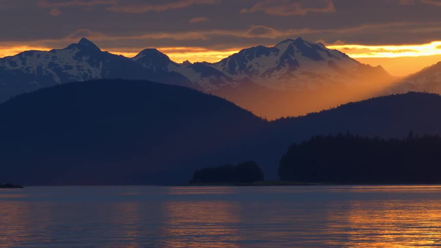 A golden sunset illuminates snow-capped mountains behind a tranquil lake