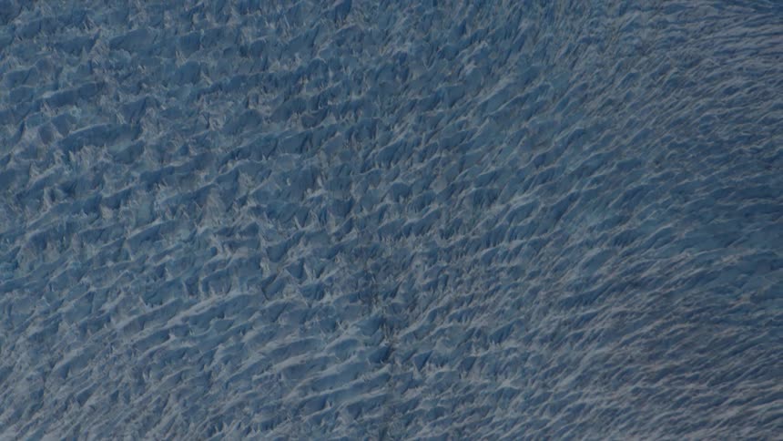 A slow, breathtaking aerial pull-out shot reveals the immense scale and intricate patterns of a vast ice glacier