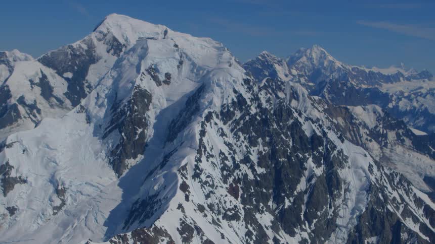 Slow aerial pullback revealing a vast and majestic snow-covered mountain range against a clear blue sky