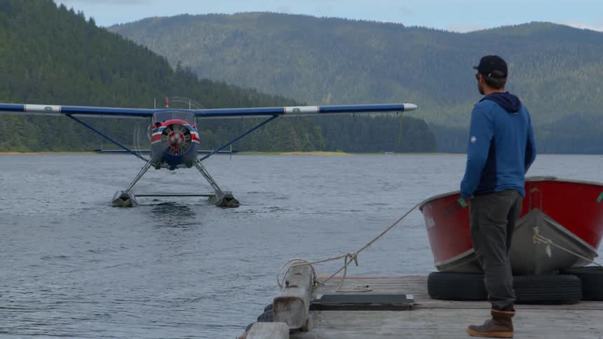 A man stands on a wooden dock and watches a seaplane taxi across a remote lake surrounded by forested mountains
