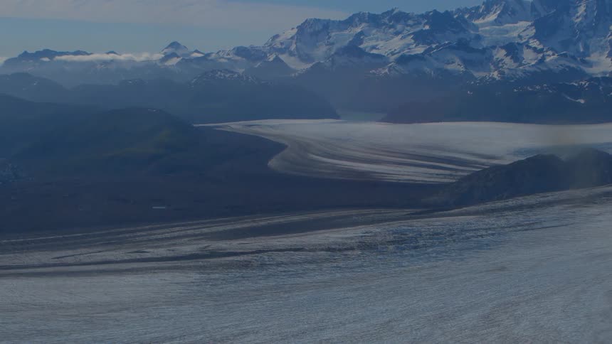 A breathtaking wide aerial shot shows a massive glacier flowing through a remote valley with a snow-capped mountain range in the background