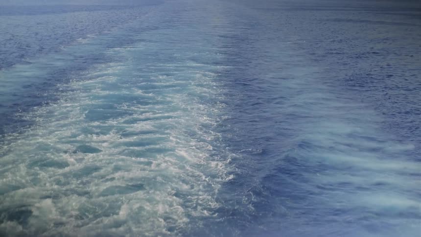 A view from the back of a boat shows the foamy white wake trailing through the deep blue ocean