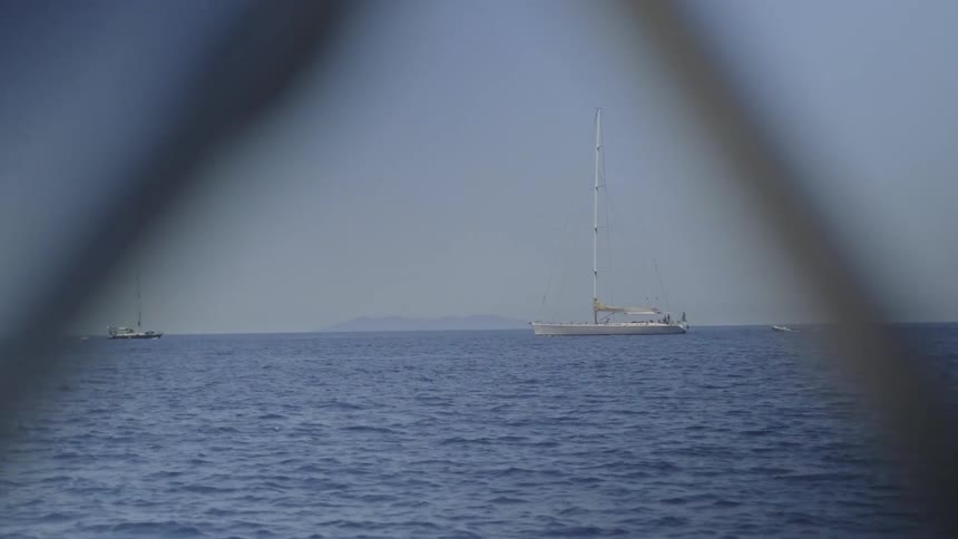 Sailboats and a motorboat peacefully float on the calm blue sea with a distant island on the horizon