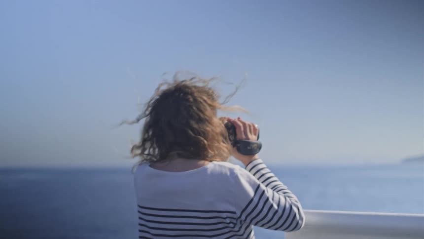 A woman with windblown hair films the vast blue ocean with a retro camcorder from the deck of a moving boat