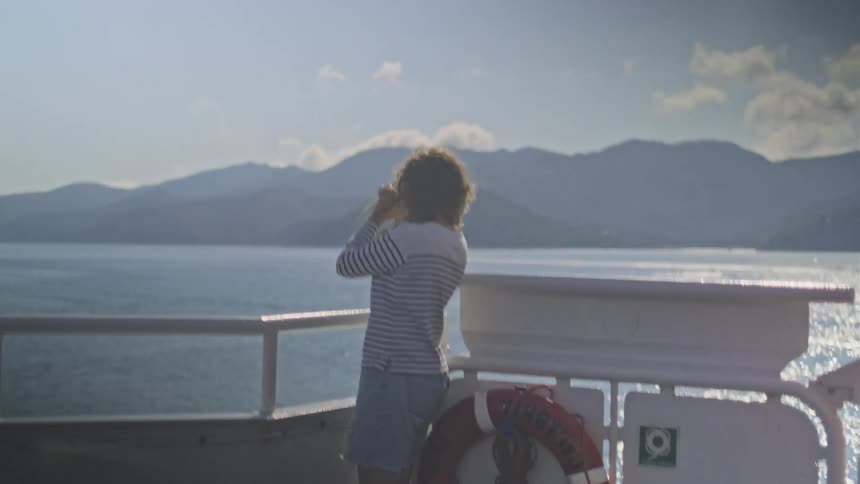 A curly-haired boy in a striped shirt stands on a ferry deck, exploring the distant mountains with a small telescope
