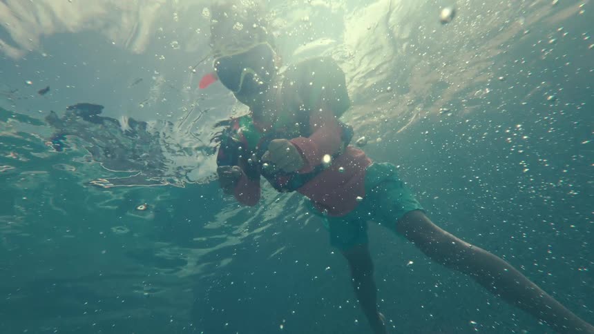An underwater slow-motion shot shows a young boy in a snorkel mask and floaties swimming in a pool with bubbles rising around him