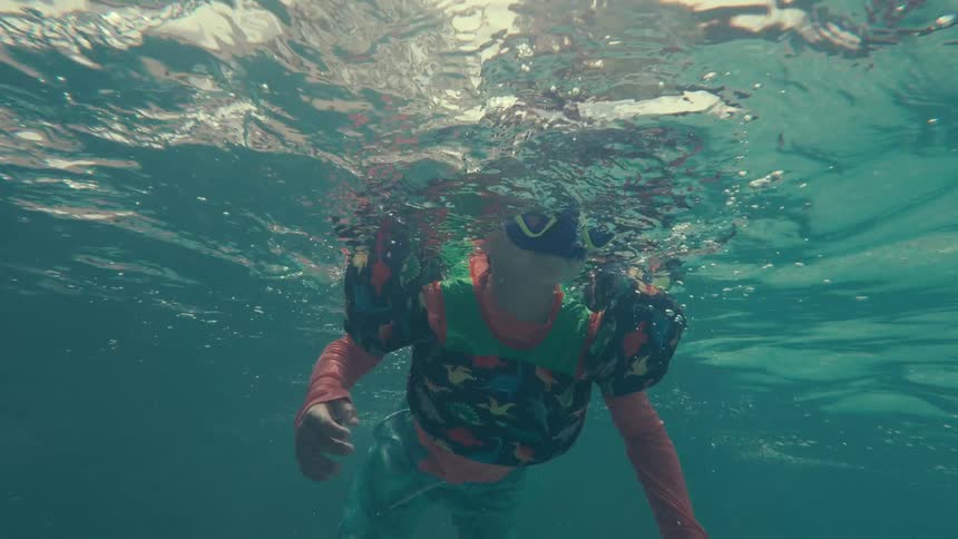 A slow motion underwater shot shows a little boy in goggles and floaties happily swimming and giving a thumbs up