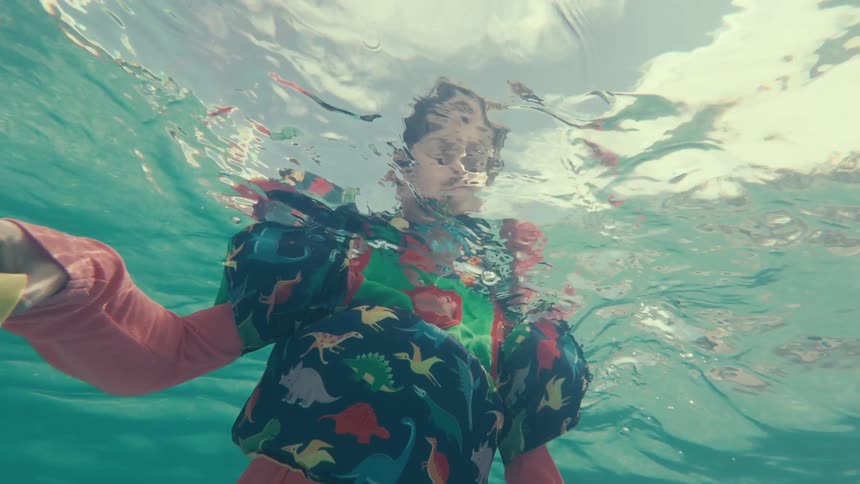 An underwater slow-motion shot looks up at a young boy in a life vest floating on the surface of the clear ocean