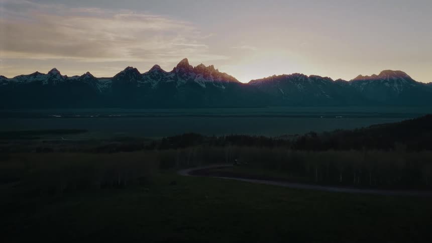 An aerial view shows a lone camper van on a winding road with a stunning sunset behind the majestic Grand Teton mountains